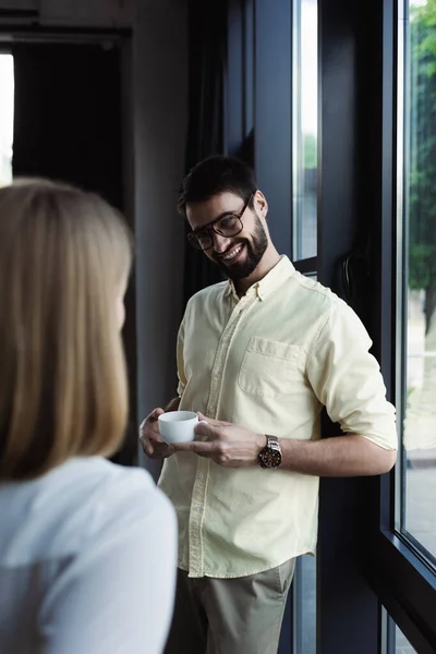 Усміхнений бізнесмен тримає чашку кави поблизу розмитого інтерну в офісі — стокове фото