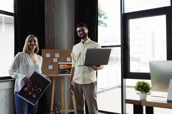 Smiling manager holding laptop and looking at camera near intern with charts in office — Stock Photo