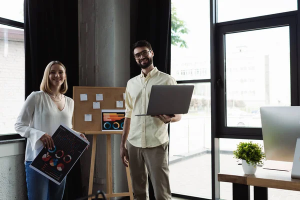 Gestionnaire souriant tenant un ordinateur portable et regardant la caméra près du stagiaire avec des graphiques dans le bureau — Photo de stock