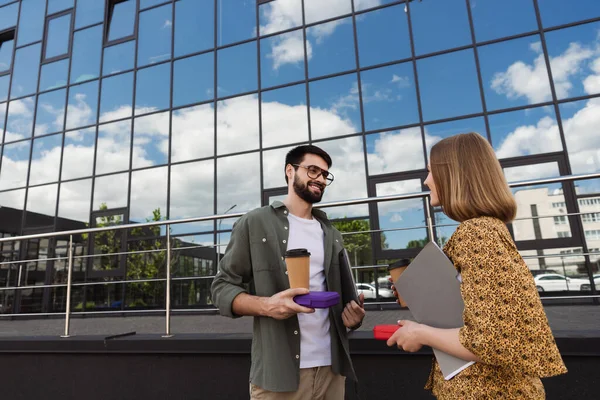 Hombre de negocios sonriente sosteniendo lonchera y café para acercarse a colega con carpeta de papel al aire libre - foto de stock