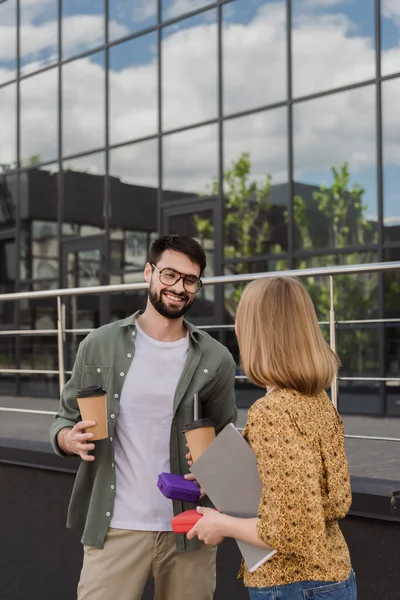 Улыбающийся бизнесмен держит коробку с обедом и кофе, чтобы пойти, разговаривая с коллегой с бумажной папкой на открытом воздухе — стоковое фото