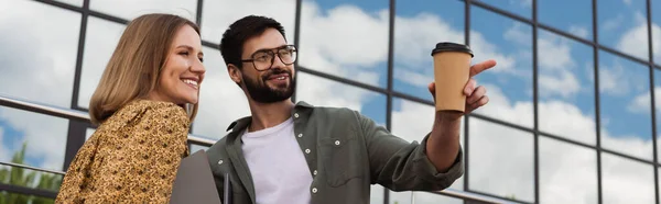 Positiver Geschäftsmann hält Coffee to go in der Hand und zeigt mit dem Finger auf Kollegen im Freien, Banner — Stockfoto
