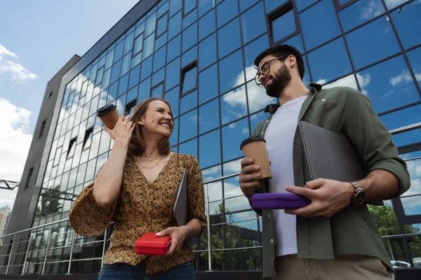 Low angle view of smiling businesswoman holding coffee to go and lunch box near colleague with laptop outdoors — Stock Photo
