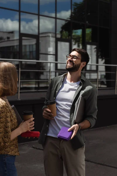 Alegre empresario sosteniendo portátil y lonchera cerca de colega con café para salir al aire libre - foto de stock