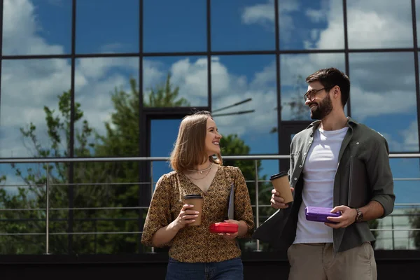 Позитивные деловые люди разговаривают, держа в руках обеденные коробки и кофе, чтобы выйти на улицу — стоковое фото