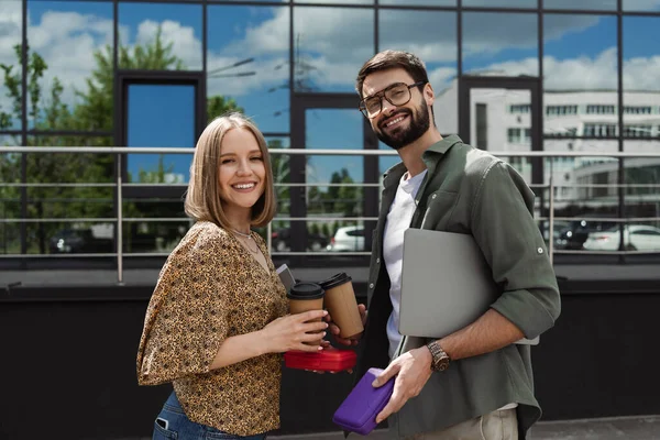 Smiling business people with laptop and lunch boxes looking at camera on urban street — Stock Photo