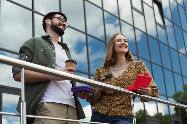 Positivi uomini d'affari che tengono caffè da asporto e scatole da pranzo vicino all'edificio sfocato all'aperto — Foto stock