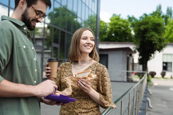 Femme d'affaires souriante tenant un sandwich et un café à emporter près d'un collègue dans une rue urbaine — Photo de stock