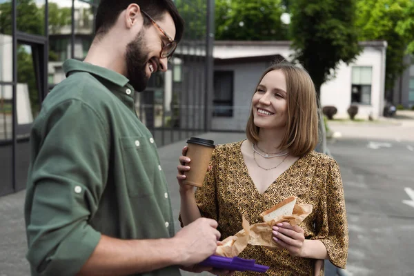 Empresária sorridente segurando sanduíche e café para ir perto colega borrado com lancheira ao ar livre — Fotografia de Stock