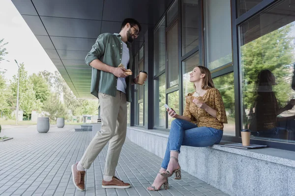 Geschäftsfrau mit Smartphone und Sandwich im Gespräch mit Kollegin, die Kaffee trinkt, um im Freien zu bauen — Stockfoto