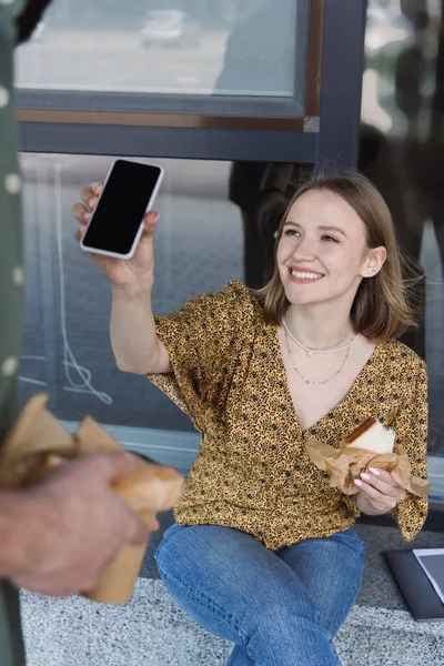 Усміхнена бізнес-леді тримає сендвіч і смартфон біля колеги на міській вулиці — стокове фото