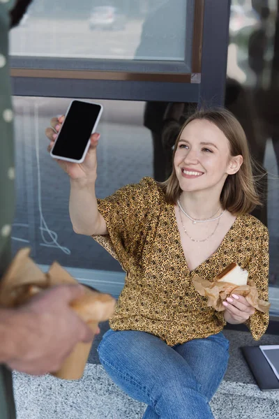 Smiling businesswoman holding sandwich and smartphone near colleague on urban street — Stock Photo
