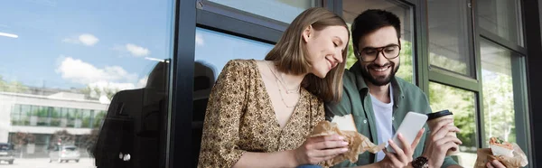 Businesswoman holding sandwich and smartphone near colleague with coffee and building on urban street, banner — Stock Photo