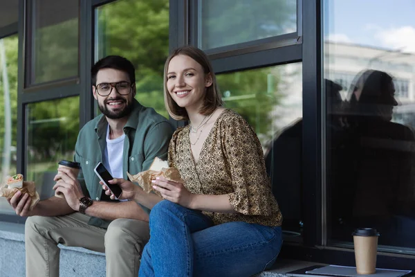 Des hommes d'affaires souriants tenant des sandwichs et un smartphone tout en regardant la caméra près du bâtiment à l'extérieur — Photo de stock