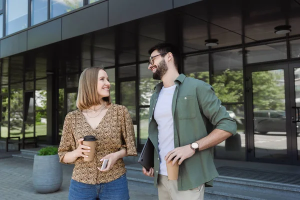 Усміхнена бізнес-леді тримає каву і мобільний телефон біля колеги з ноутбуком на відкритому повітрі — стокове фото