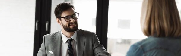 Hombre de negocios positivo en ropa formal mirando a la mujer borrosa durante la entrevista de trabajo en la oficina, pancarta - foto de stock