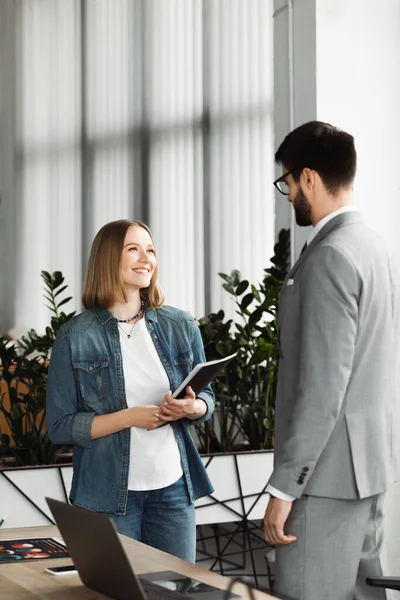 Sonriente solicitante de empleo sosteniendo carpeta de papel cerca de hombre de negocios durante la entrevista en la oficina - foto de stock
