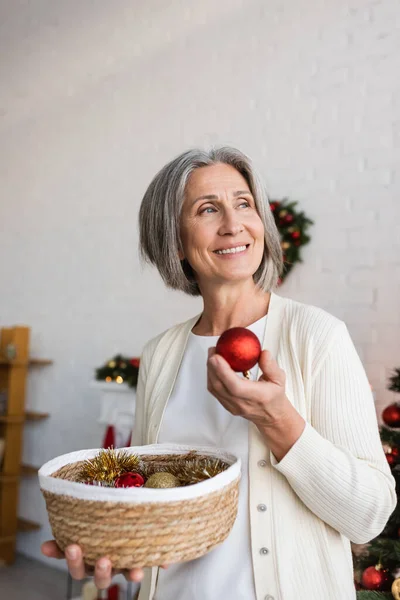 Happy middle aged woman with grey hair holding wicker basket and red christmas ball — Stock Photo