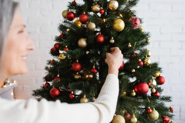 Verschwommene und fröhliche Frau mittleren Alters mit grauen Haaren, die den Weihnachtsbaum schmückt — Stockfoto