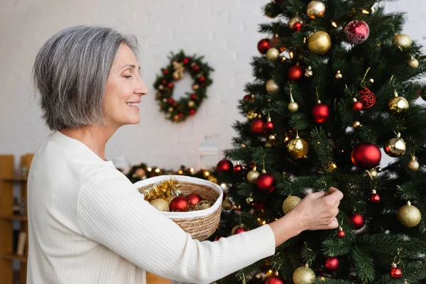 Mujer de mediana edad feliz con pelo gris sosteniendo canasta de mimbre y decorando el árbol de Navidad - foto de stock