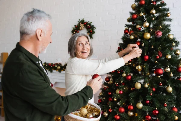 Mujer de mediana edad feliz con pelo gris decorando el árbol de Navidad y mirando al marido sosteniendo la cesta de mimbre - foto de stock