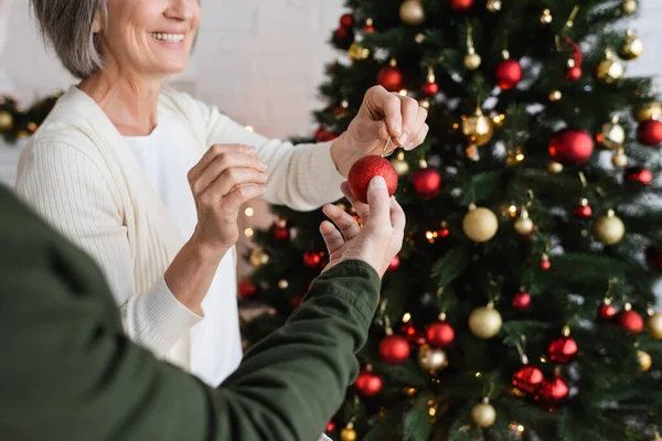 Vista parcial del hombre de mediana edad dando juguete de navidad a la esposa feliz al decorar el pino en casa - foto de stock