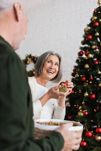 Alegre mulher de meia idade com cabelos grisalhos decorando árvore de natal perto do marido — Fotografia de Stock