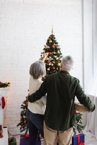 Vista trasera de pareja de mediana edad decorando el árbol de Navidad en casa - foto de stock
