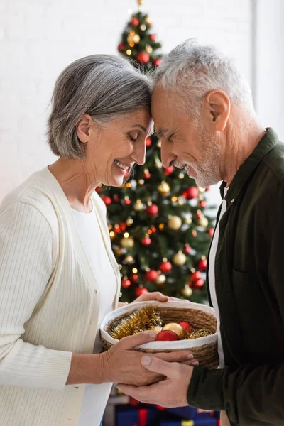 Smiling middle aged couple with closed eyes holding wicker basket near decorated christmas tree — Stock Photo