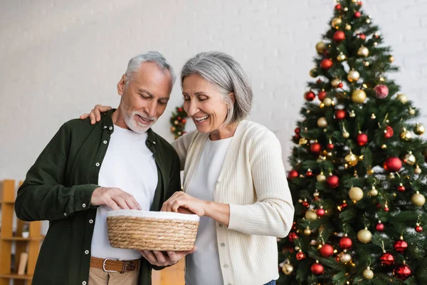 Smiling middle aged couple looking at wicker basket near decorated christmas tree — Stock Photo