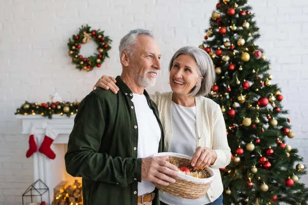 Smiling middle aged couple holding wicker basket with baubles near decorated pine tree — Stock Photo