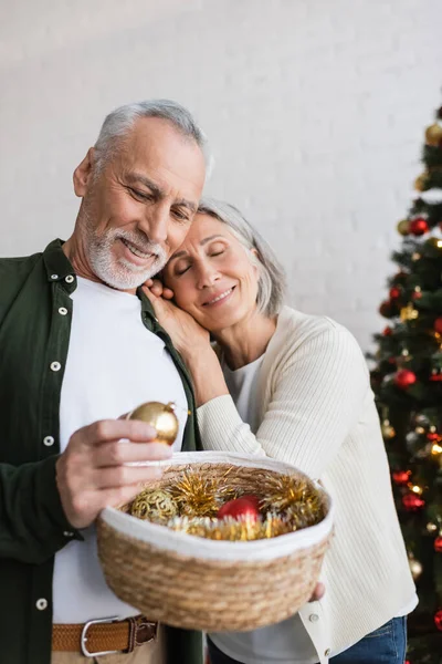 Sorrindo mulher de meia-idade apoiando-se no marido com cesta de vime perto da árvore de natal — Fotografia de Stock