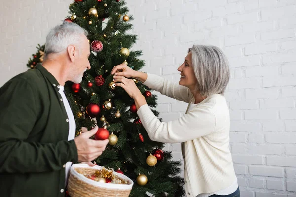Alegre casal de meia idade segurando bugigangas enquanto decora árvore de Natal — Fotografia de Stock