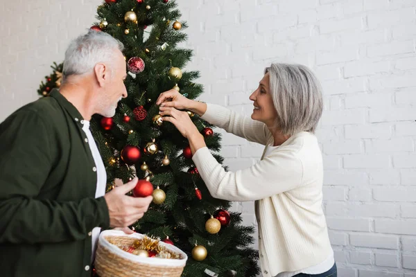 Cheerful middle aged couple holding baubles while decorating christmas tree — Stock Photo
