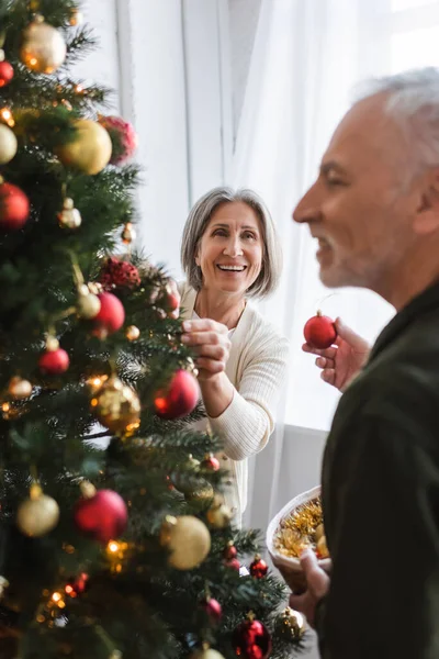 Cheerful middle aged woman decorating christmas tree and looking at bearded husband — Stock Photo
