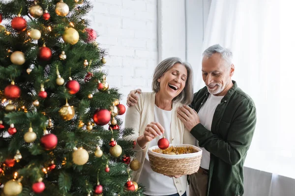 Fröhliche Frau mittleren Alters lacht, während sie Christbaumkugel in der Nähe von Mann und Weihnachtsbaum hält — Stockfoto