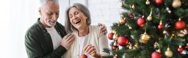 Веселая женщина средних лет смеется, держа безделушку возле мужа и рождественскую елку, плакат — стоковое фото