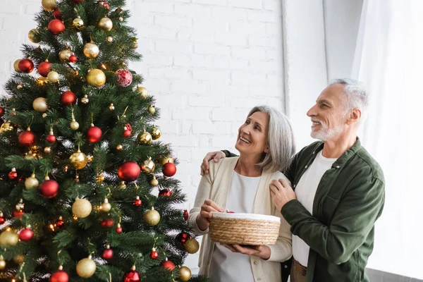 Весёлые муж и жена средних лет смотрят на украшенную елку — стоковое фото