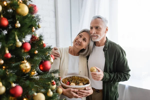 Cheerful middle aged husband hugging wife and looking at decorated christmas tree — Stock Photo
