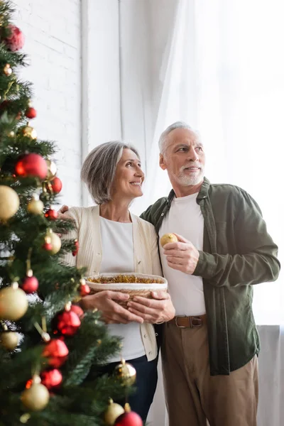 Joyeux mari d'âge moyen étreignant femme et regardant loin près décoré arbre de Noël — Photo de stock