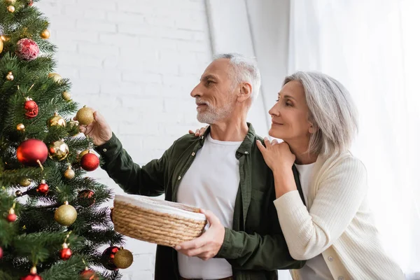 Fröhliche Frau mittleren Alters umarmt Ehemann schmückt Weihnachtsbaum — Stockfoto