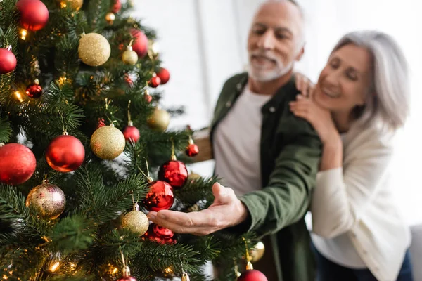 Smiling middle aged woman hugging blurred husband decorating christmas tree — Stock Photo