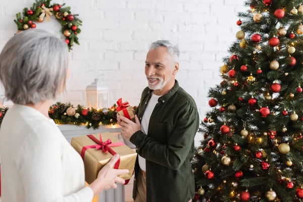 Sonriente hombre de mediana edad sosteniendo presente y mirando a la esposa cerca del árbol de Navidad - foto de stock