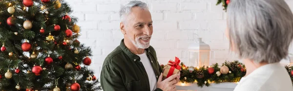 Усміхнений чоловік середнього віку тримає подарунок і дивиться на дружину біля ялинки, банер — стокове фото