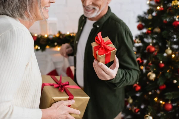 Обрізаний вид веселого чоловіка середнього віку, що тримає подарунок біля дружини та ялинки — стокове фото