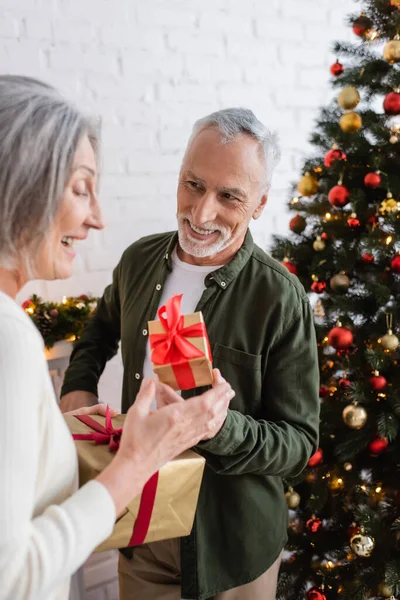 Feliz homem de meia idade segurando caixa de presente e olhando para a esposa perto da árvore de natal — Fotografia de Stock