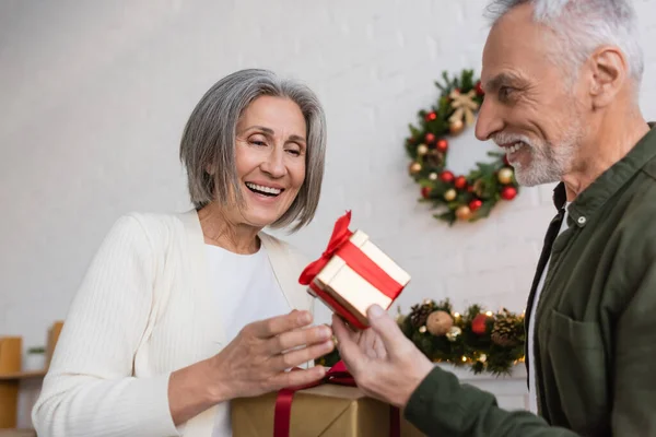 Feliz hombre de mediana edad sosteniendo regalo de Navidad cerca de esposa alegre - foto de stock