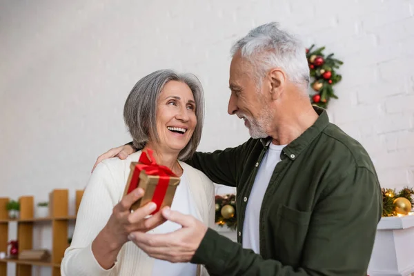 Sonriente madura mujer celebración navidad presente cerca alegre marido - foto de stock