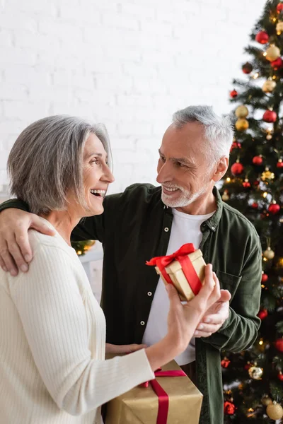 Усміхнена зріла жінка тримає різдвяний подарунок біля веселого чоловіка і прикрашена ялиною — стокове фото