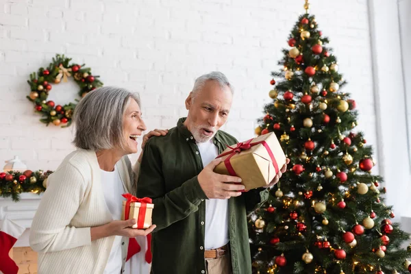 Souriant femme mûre tenant cadeau de Noël et regardant mari surpris près de sapin décoré — Photo de stock